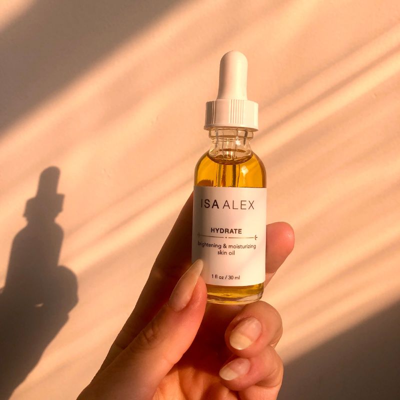 Hand holding a bottle of ISA ALEX skincare oil with a white label against a warm-toned background
