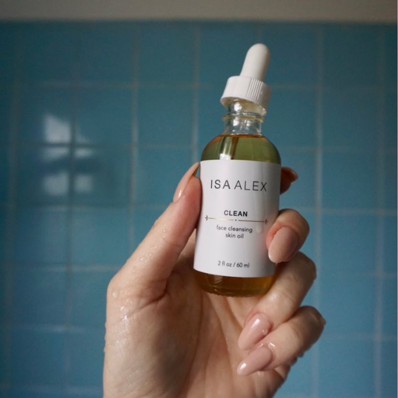 Hand holding a bottle of Isa Alek skincare oil against a blue tiled wall.